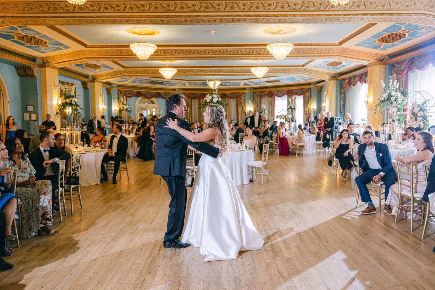 A joyous father-daughter dance in a beautifully decorated ballroom filled with guests enjoying the celebration.