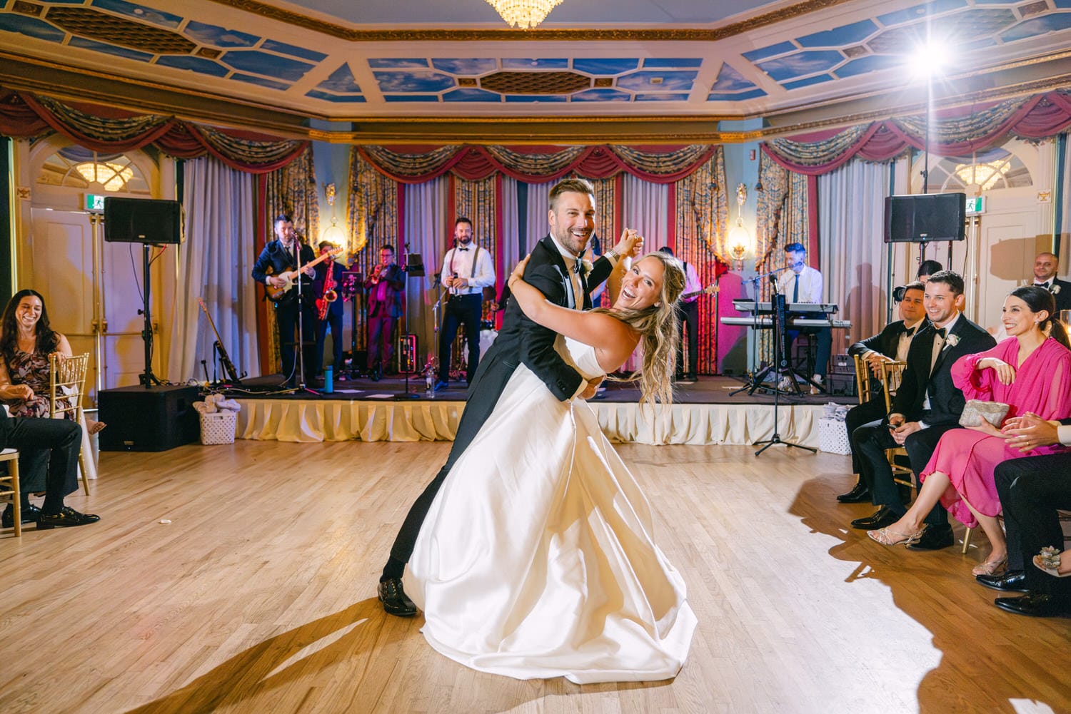 A joyful couple dancing at their wedding reception, surrounded by friends and a live band in an elegantly decorated venue.