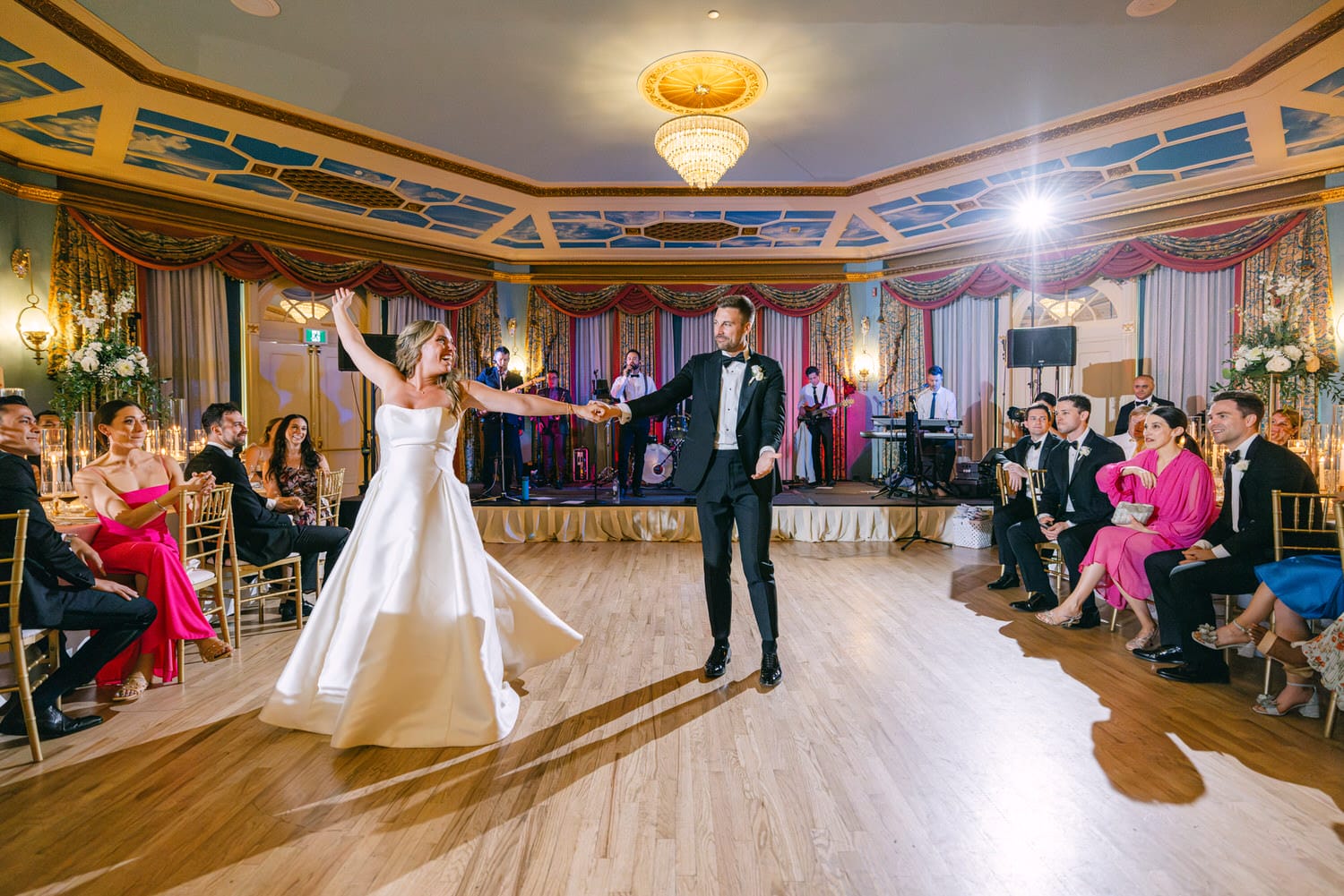 A joyful wedding couple dancing, surrounded by guests seated at elegantly set tables, with a live band performing in the background.