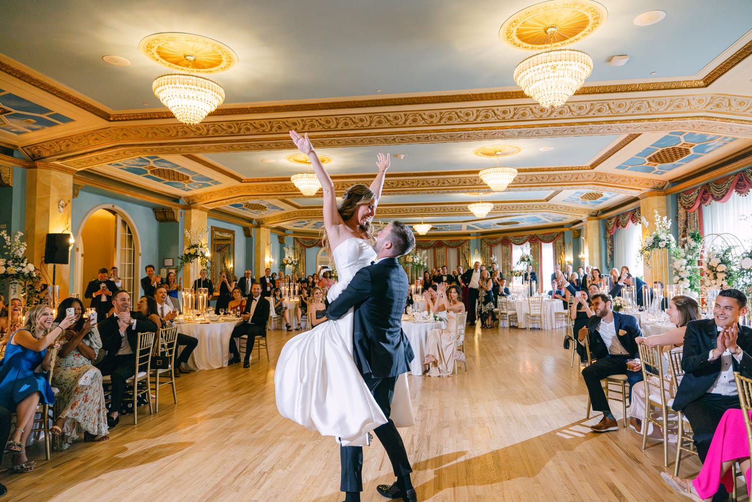 A joyful bride is lifted by her groom during their wedding reception, surrounded by applauding guests and an elegant venue.