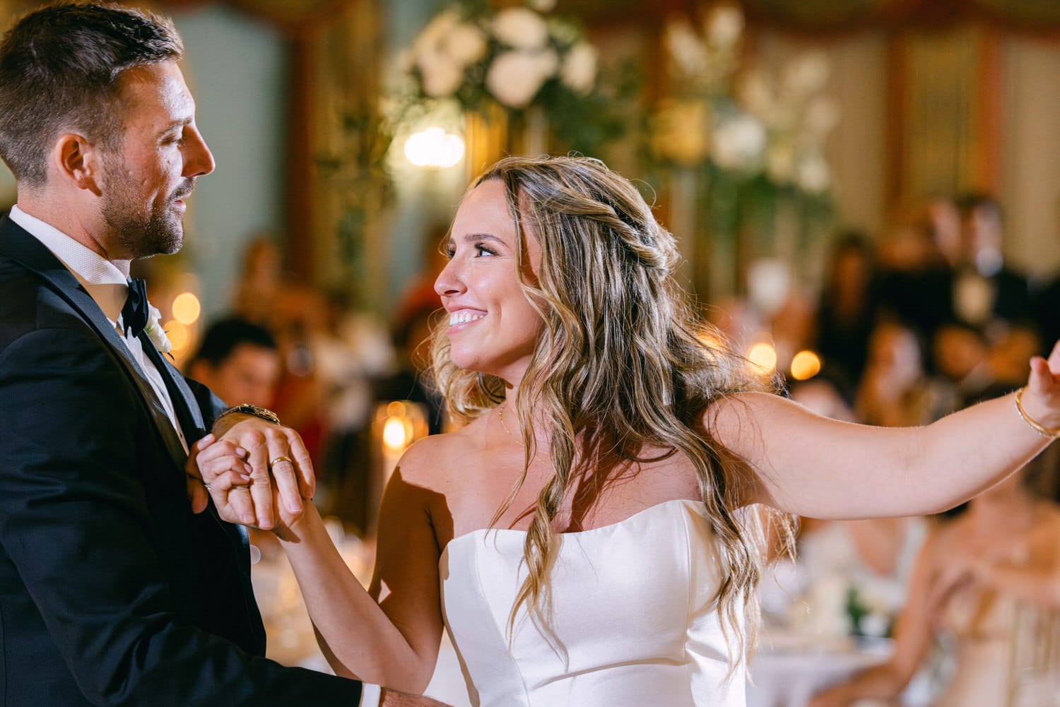 A bride and groom share a joyful moment during their first dance at an elegant wedding reception, surrounded by warm candlelight and guests.