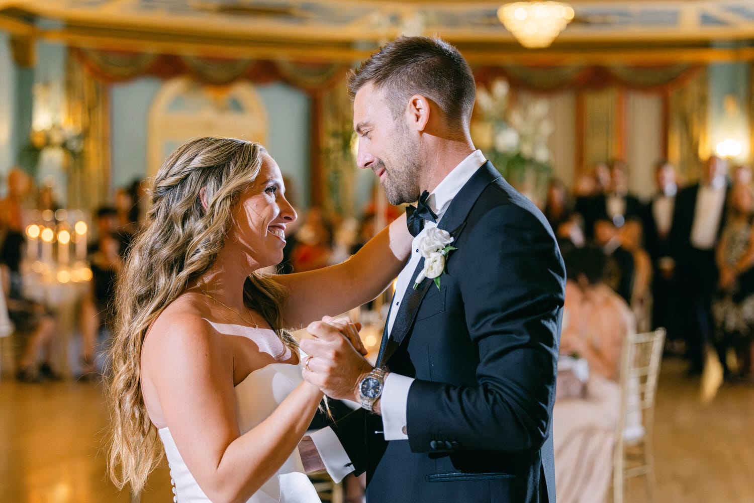 A bride and groom share a romantic moment during their first dance at a wedding reception, surrounded by elegantly dressed guests in a beautifully decorated venue.