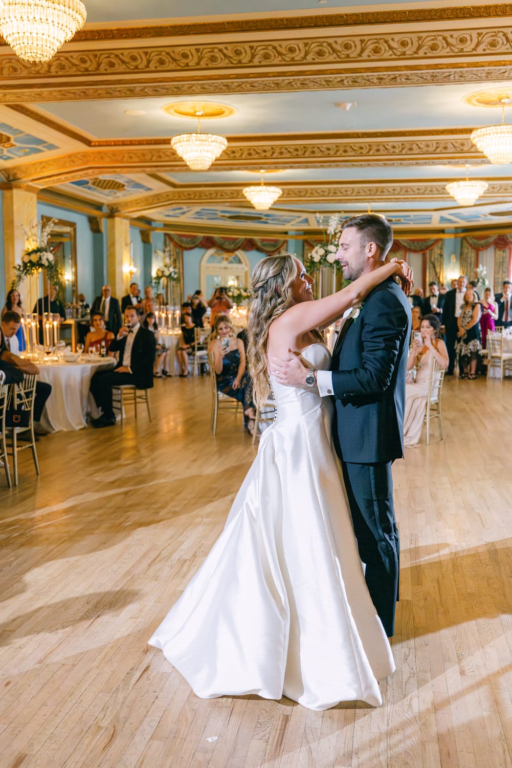 A newlywed couple shares a romantic first dance in an ornate ballroom surrounded by seated guests and candlelight.