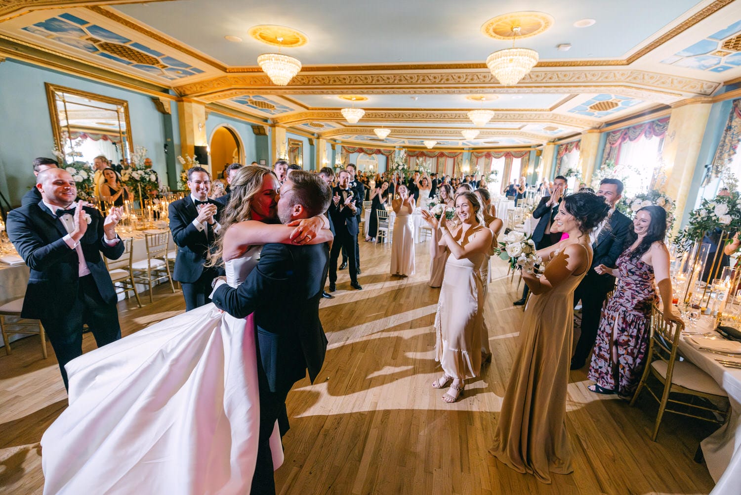 A bride and groom share a romantic embrace while celebrating with guests in an elegantly decorated reception hall.