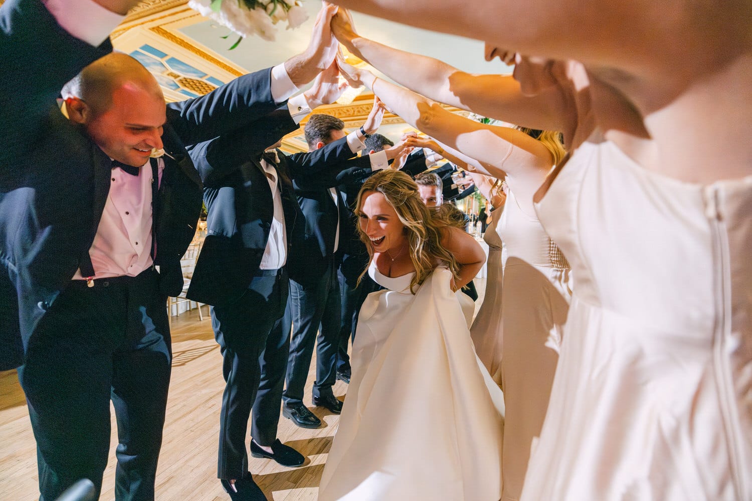 A joyful bride laughing and dancing under an arch of raised arms during her wedding celebration, surrounded by guests in formal attire.