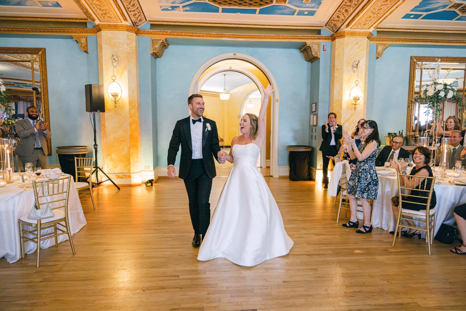 A joyful couple enters the reception, hand in hand, under the admiration of guests in a beautifully decorated venue.