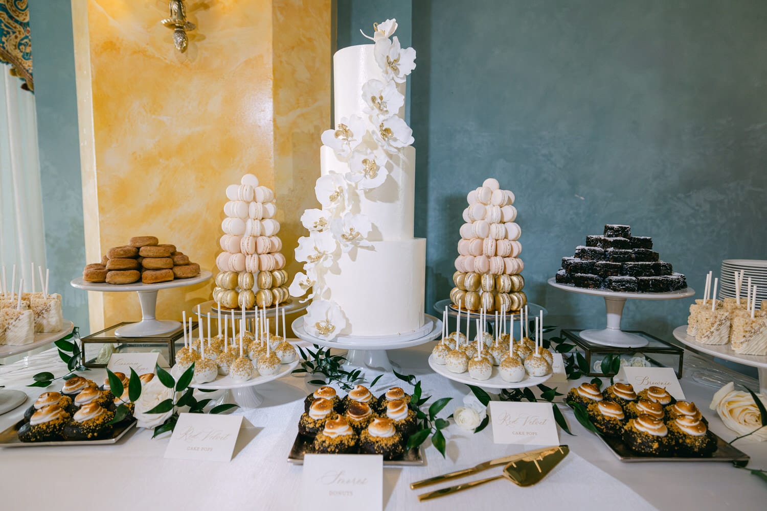 A beautifully arranged dessert table featuring a tall, white wedding cake adorned with orchids, macarons in various colors, cake pops, donuts, and other sweet treats, set against a stylish background.