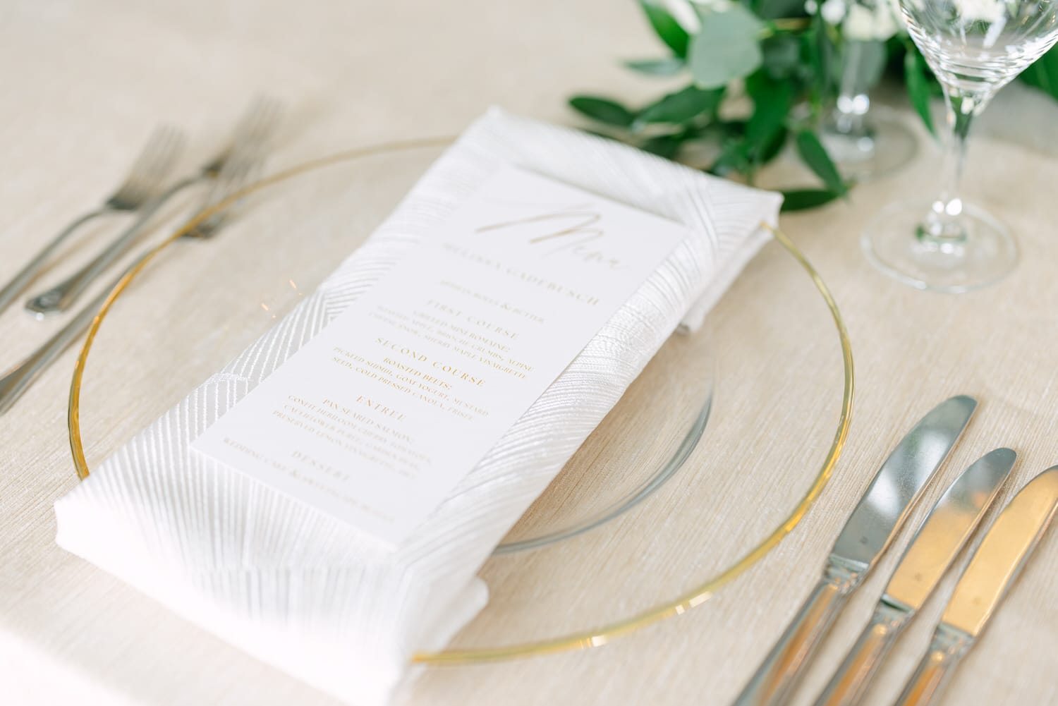 A beautifully arranged table setting featuring a menu on a decorative napkin, surrounded by glassware and silver cutlery, set against a soft beige tablecloth.