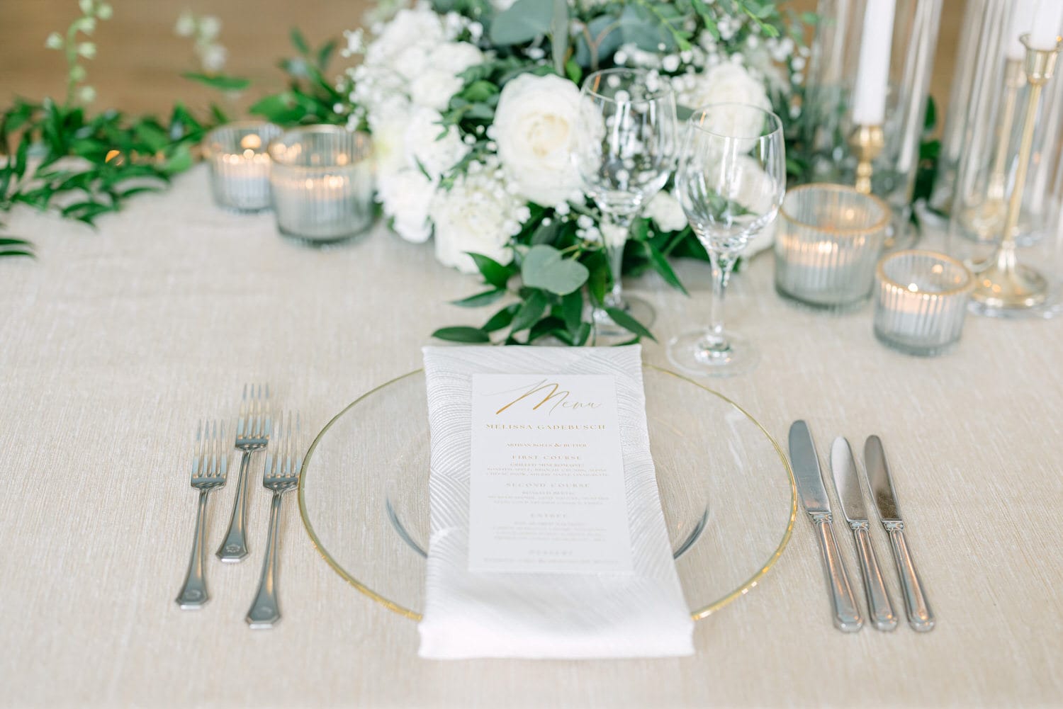 A beautifully arranged dinner table featuring a glass plate, silver cutlery, a menu card, delicate floral arrangements, and candles creating a warm ambiance.