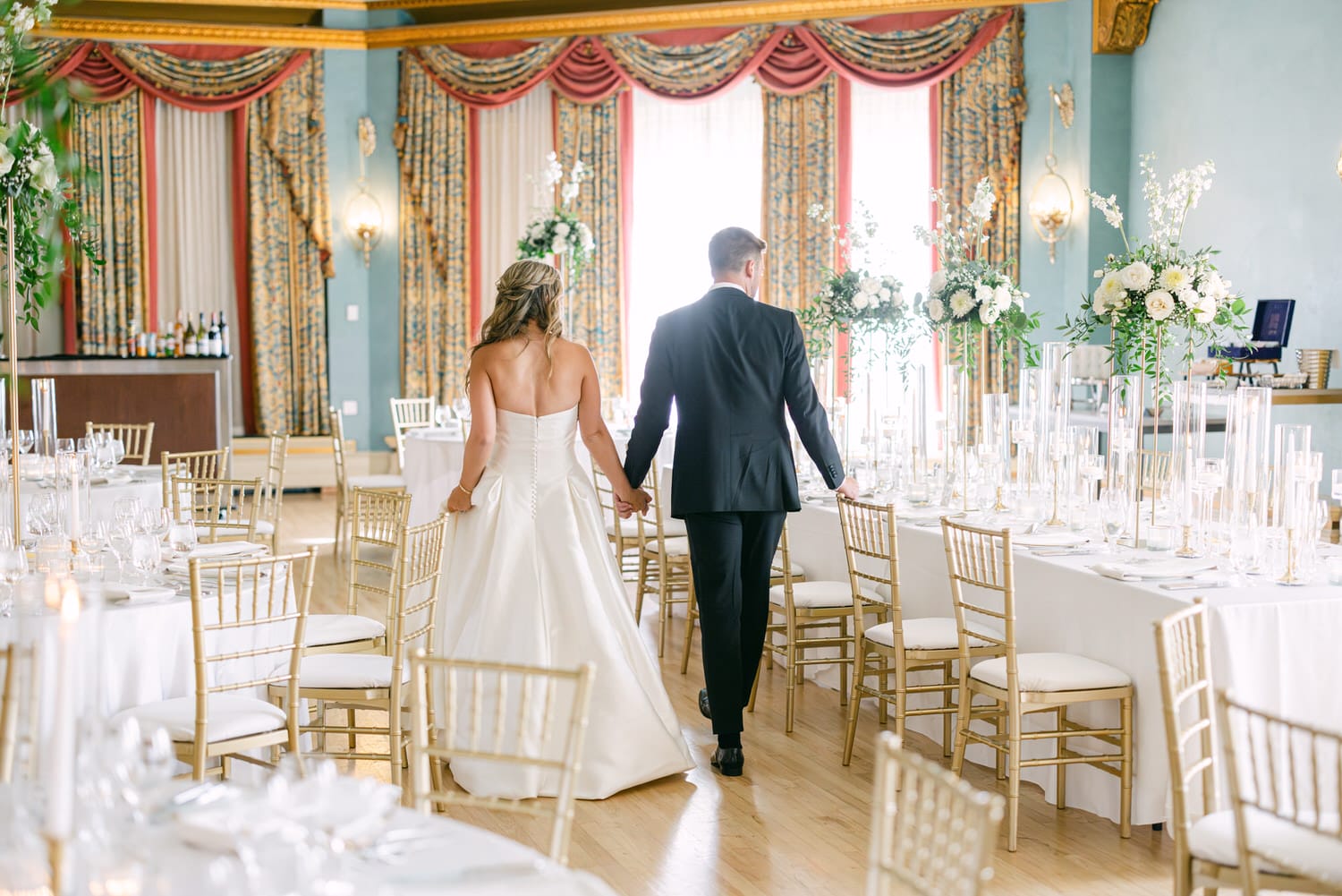 A couple walks hand in hand through a beautifully decorated dining area, featuring elegantly set tables and floral arrangements.