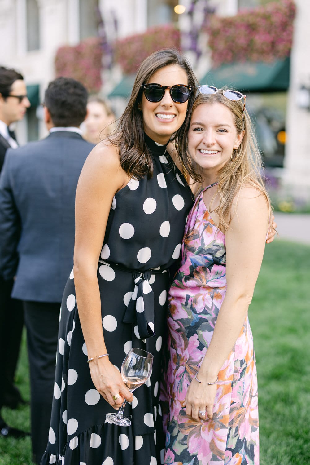 Two women posing together with smiles at an outdoor event, one in a black and white polka dot dress holding a glass of wine and the other in a floral-patterned dress.