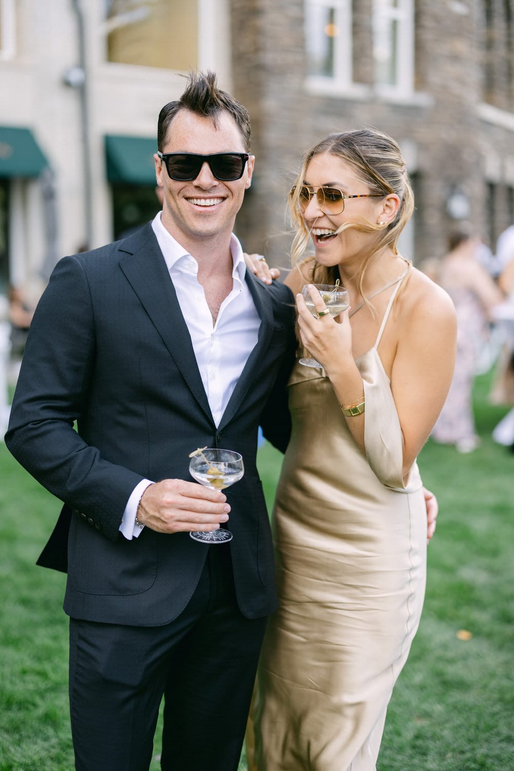 A man in a black suit and sunglasses and a woman in a gold dress enjoy drinks while laughing together at a lively outdoor gathering.