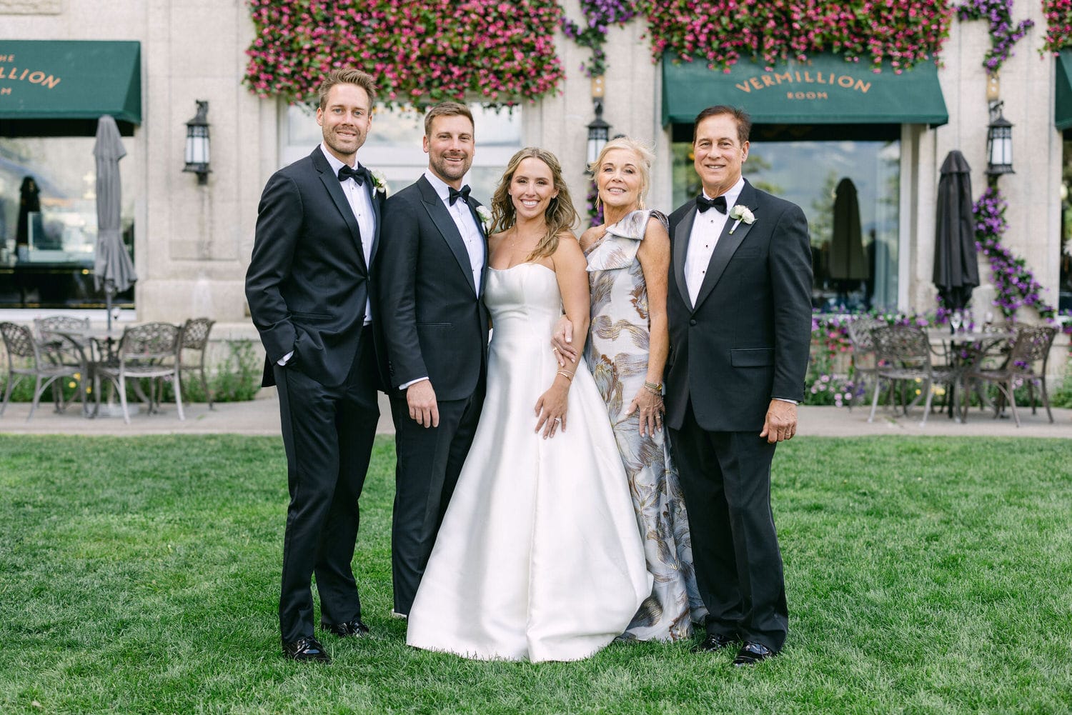 A family poses together in formal wear, outside a beautifully adorned venue with vibrant flowers and greenery.