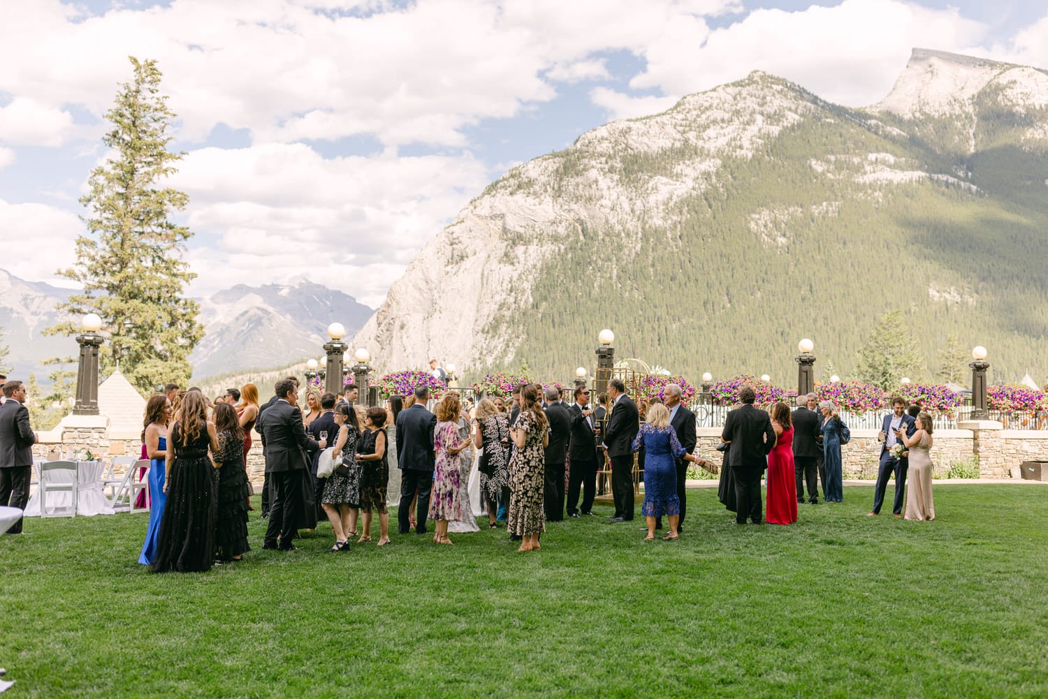 A diverse group of guests enjoying a social event on a lush green lawn, surrounded by mountains and decorative flowers.