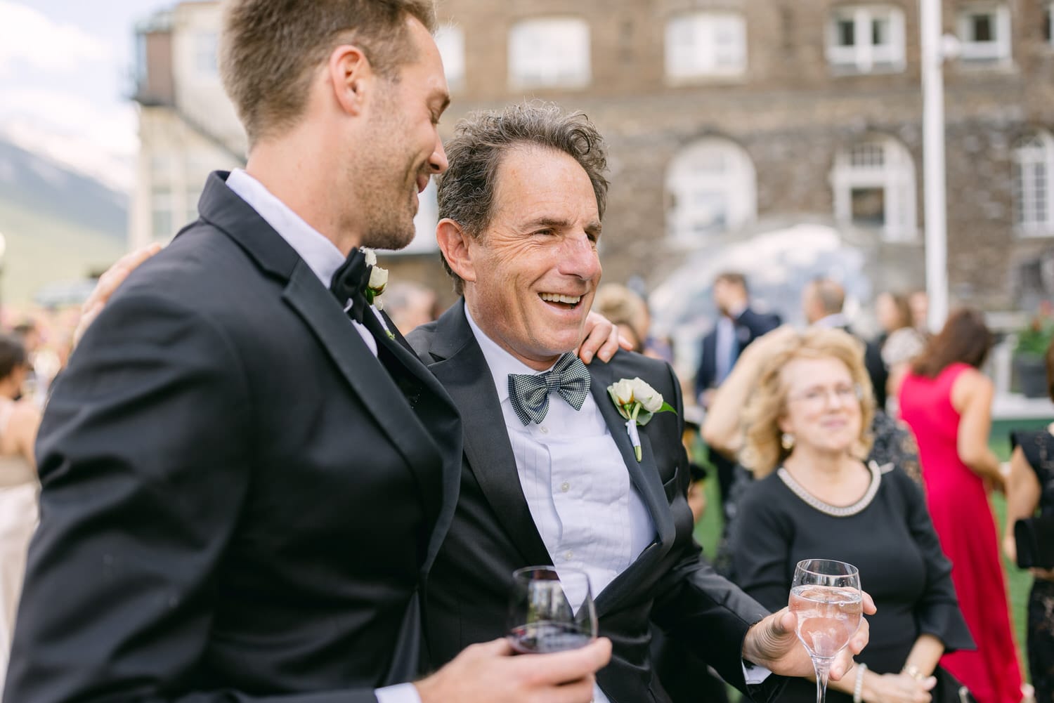 A father and son share a joyful moment during a celebration, dressed in formal attire, with lively guests in the background.