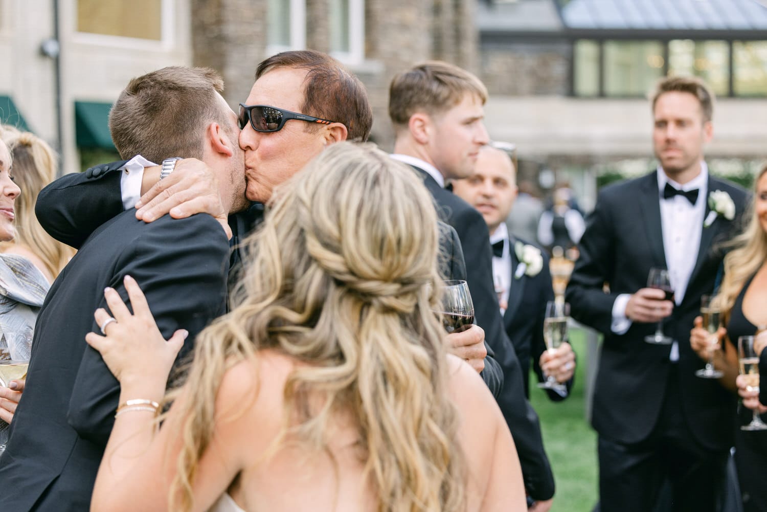 A heartfelt embrace during a wedding reception, with guests in formal attire enjoying the moment and holding drinks.