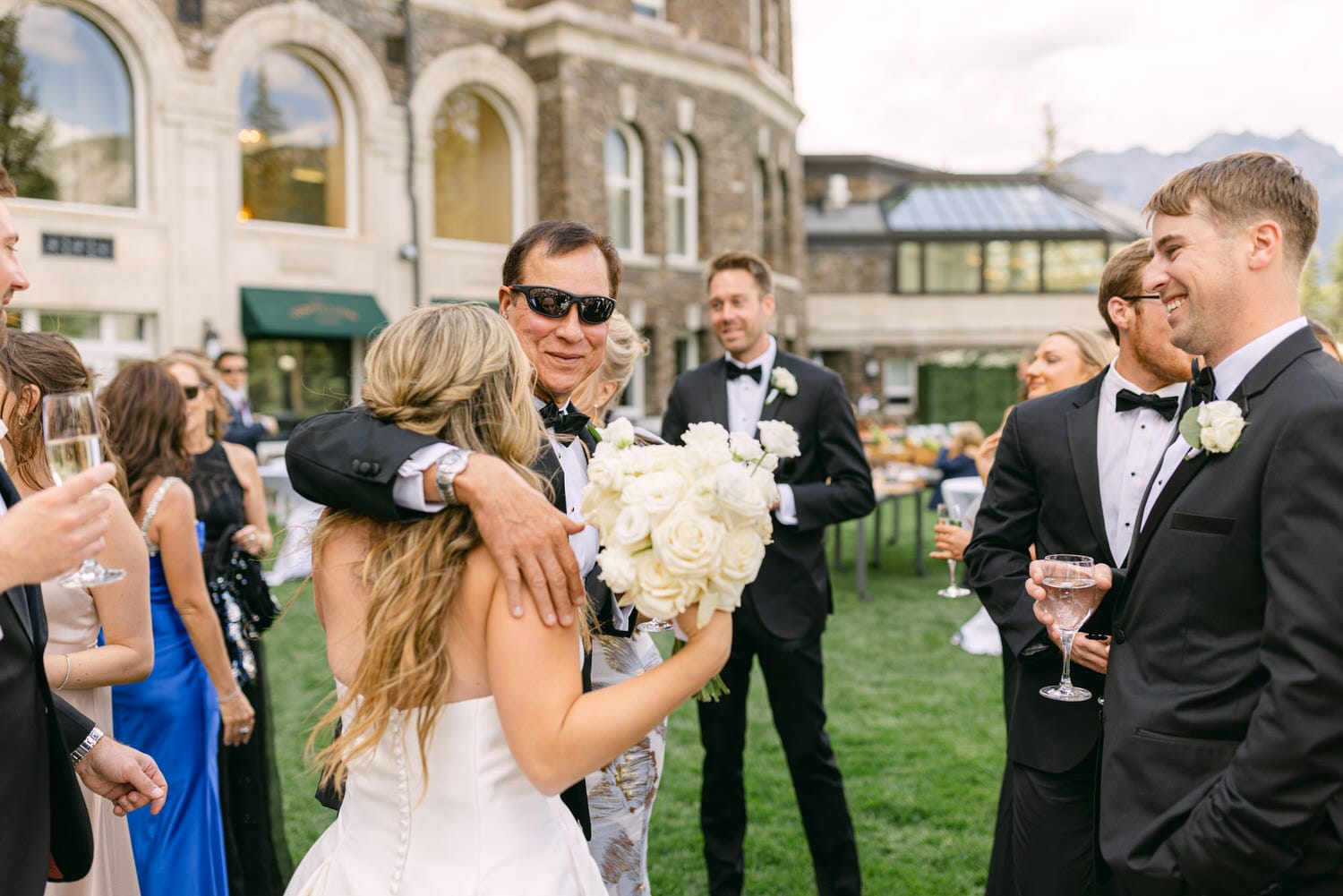 A joyful gathering featuring a bride hugging a man in sunglasses, surrounded by elegantly dressed guests enjoying a beautiful day.