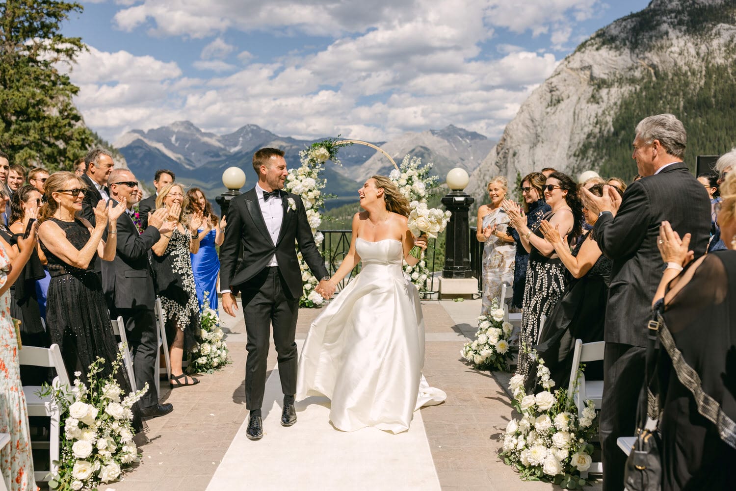 A couple joyfully exits their wedding ceremony, hand in hand, as guests applaud in a picturesque outdoor setting with mountains in the background.