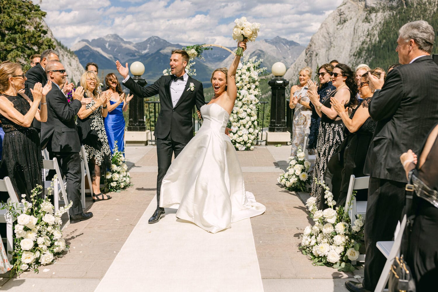 A newlywed couple exits their outdoor wedding ceremony, cheering and smiling, while guests applaud amidst a mountain backdrop adorned with floral arrangements.