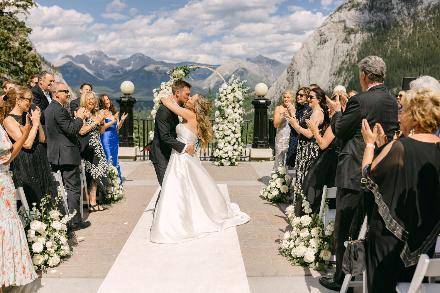 A couple shares a kiss after their wedding ceremony, surrounded by applauding guests and a stunning mountain backdrop.