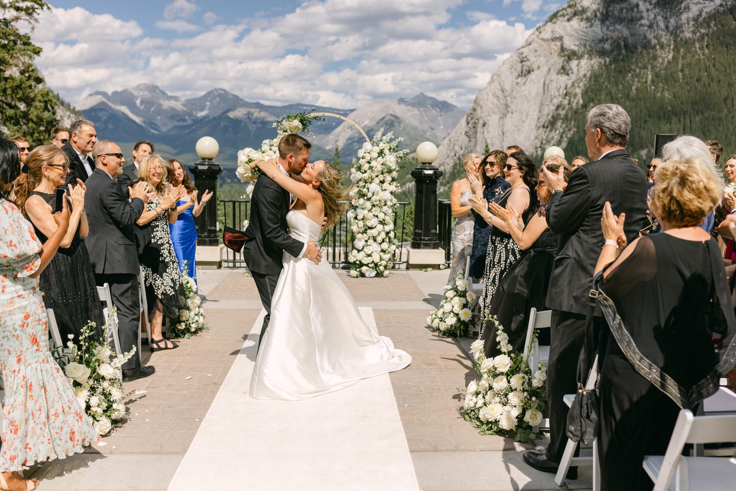 A joyful couple shares their first kiss as newlyweds in a scenic outdoor wedding surrounded by friends and family, with mountains in the background.