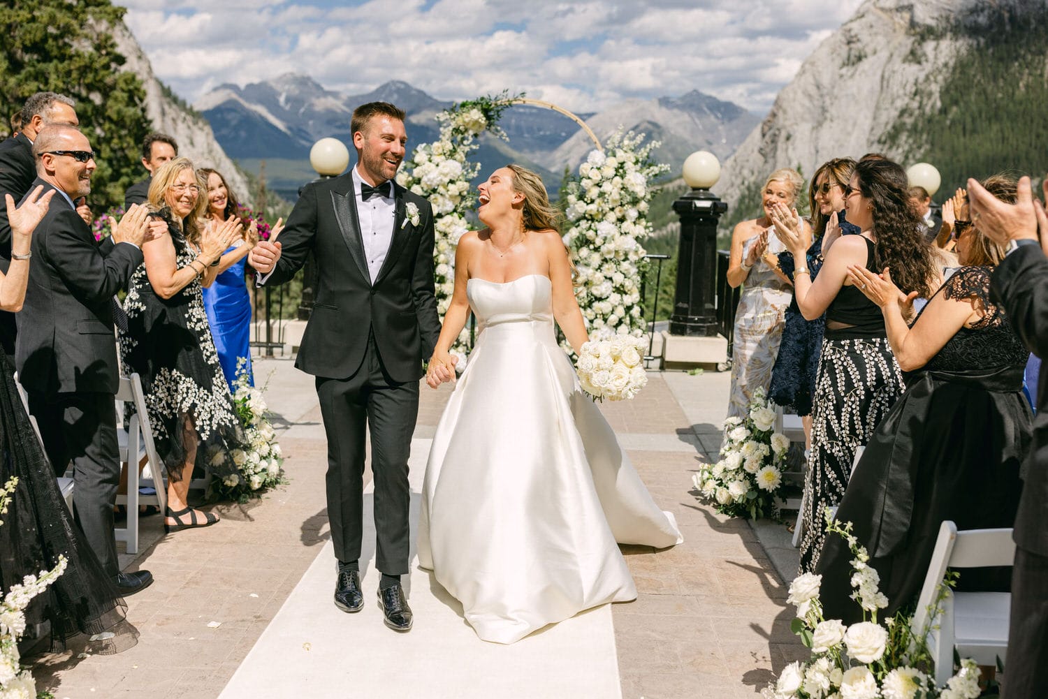 A couple joyfully exits their wedding ceremony, surrounded by applauding guests and a beautiful scenic backdrop of mountains.