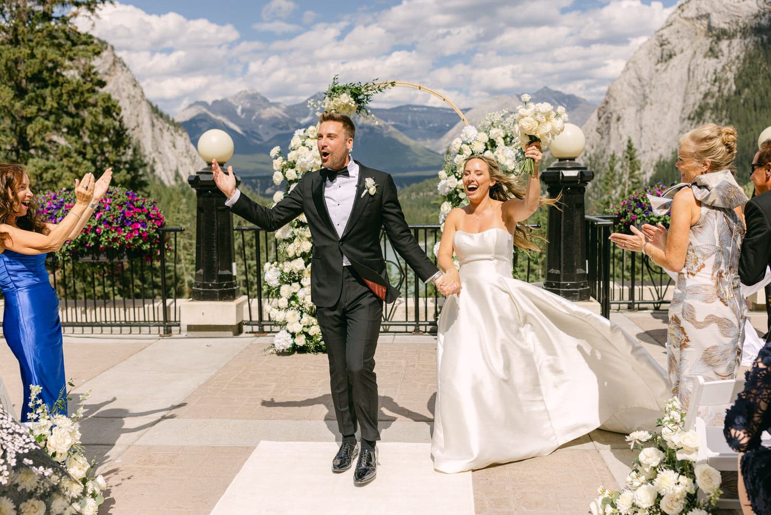 A bride and groom exuberantly celebrate their wedding against a stunning mountain backdrop, surrounded by happy guests and floral arrangements.