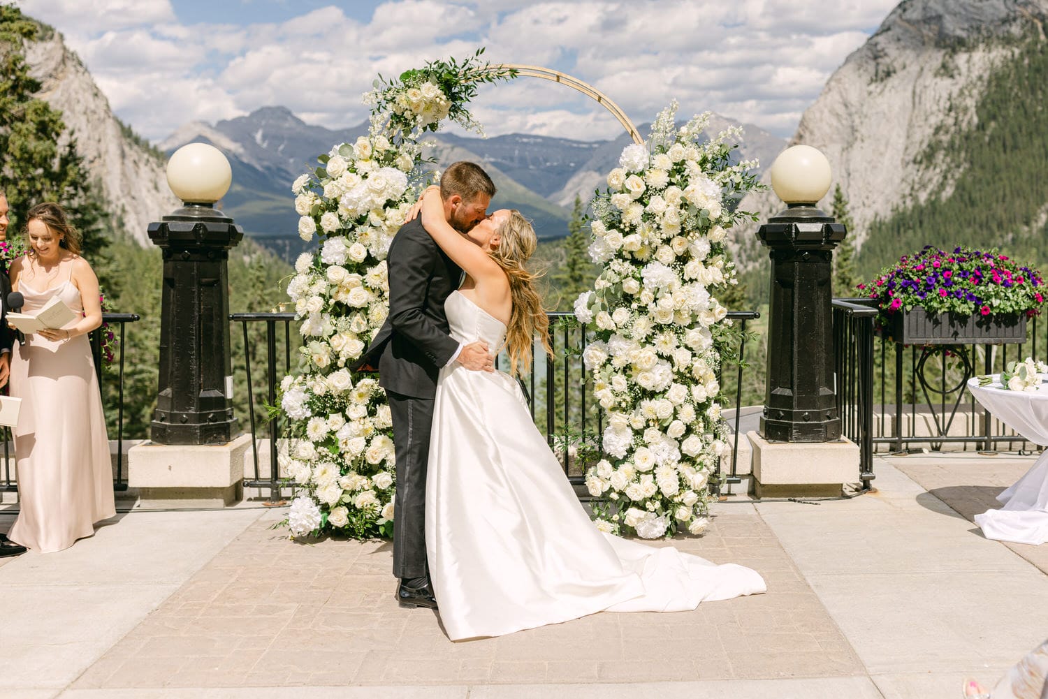 A couple shares a romantic kiss at their wedding ceremony, surrounded by lush floral decorations and majestic mountains in the background.