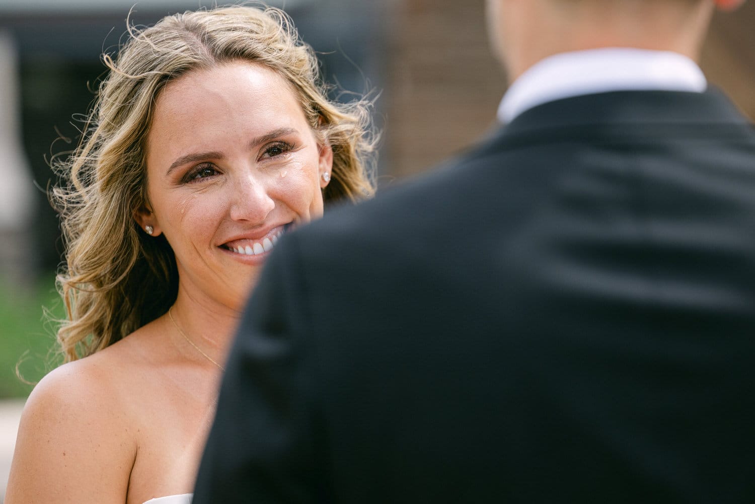 A close-up of a smiling bride with wavy hair, shedding a tear of joy while looking at her partner during a heartfelt moment.