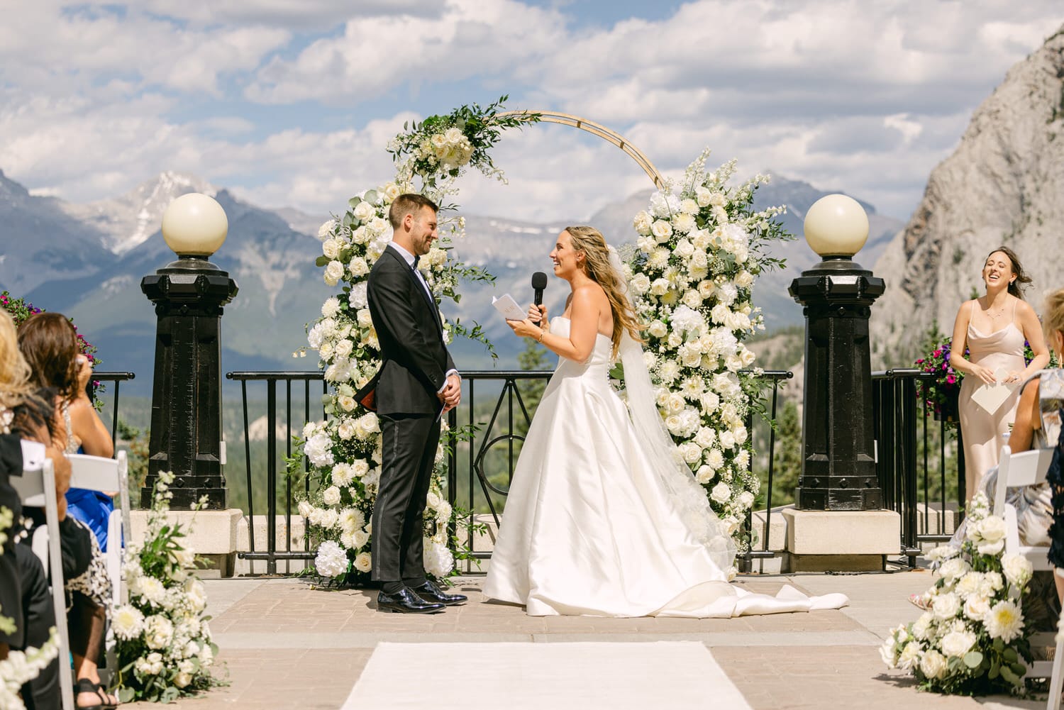 A bride and groom exchange vows during an outdoor wedding ceremony, surrounded by floral arrangements and majestic mountains under a bright blue sky.