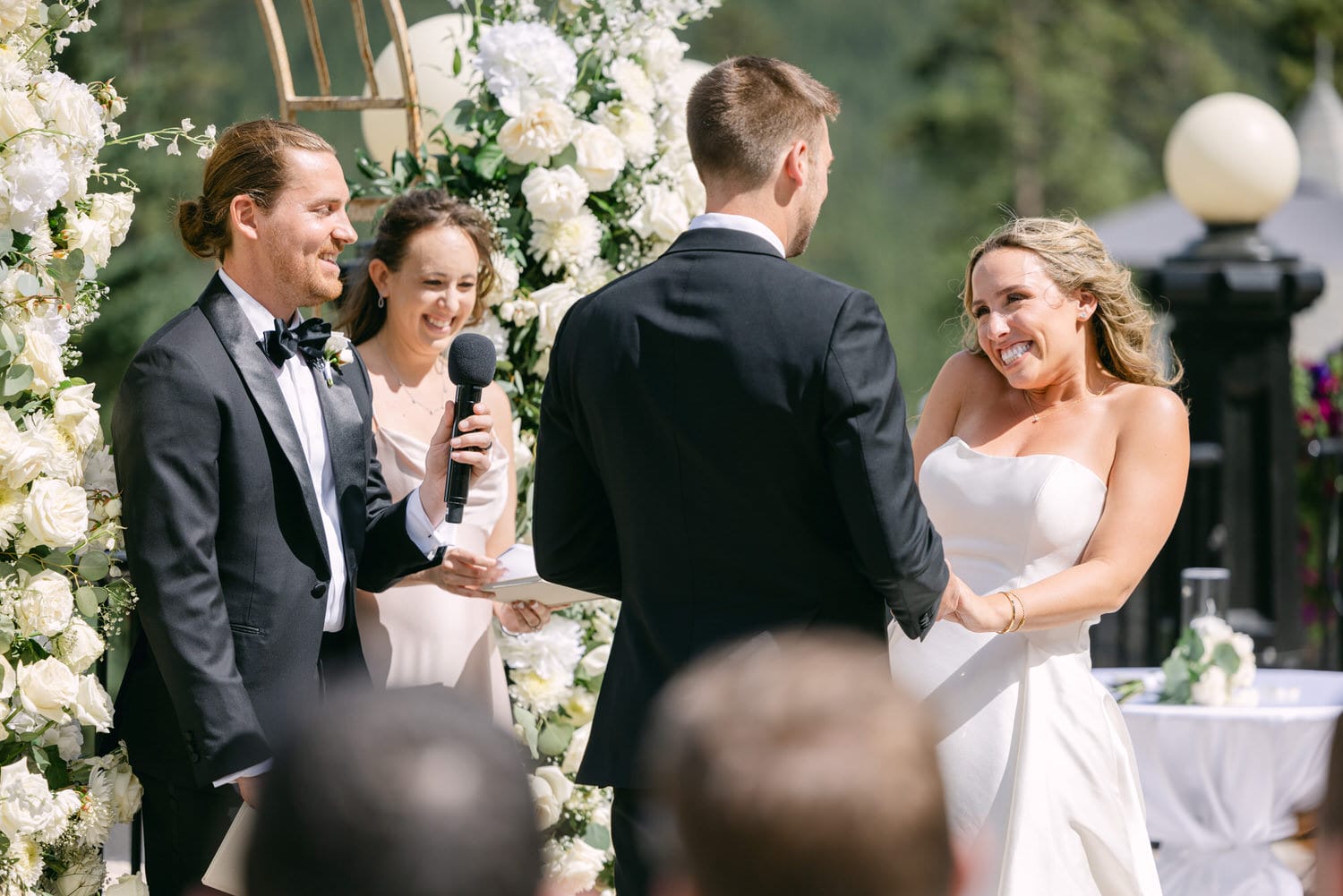 A joyful bride smiling at her groom during their outdoor wedding ceremony, surrounded by floral decorations and guests.