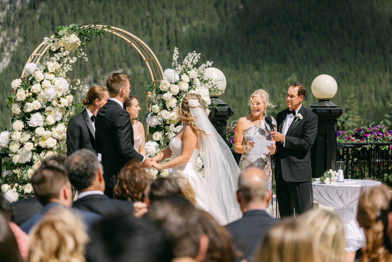 A bride and groom exchange vows in front of a floral arch, with guests seated in the foreground, amidst a picturesque mountain backdrop.