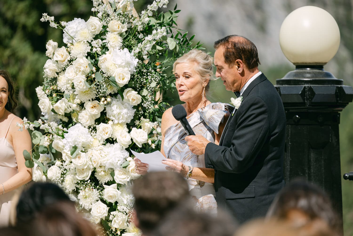 A couple shares heartfelt vows during an outdoor wedding ceremony, surrounded by a display of white flowers and attentive guests.