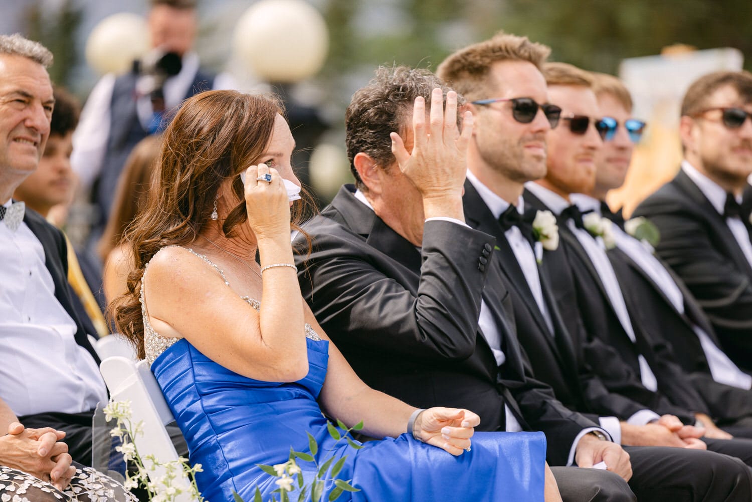 Guests experiencing heartfelt emotions during a wedding, with a woman in a blue dress wiping tears and a man beside her showing a poignant gesture.