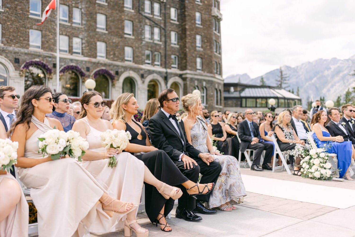 A diverse group of wedding guests seated outdoors, dressed in elegant attire, with flower bouquets and a scenic backdrop of mountains and a historic building.