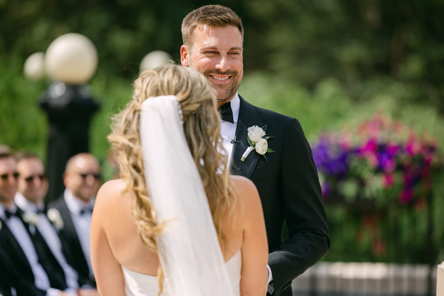 A groom smiles warmly at his bride during a wedding ceremony, surrounded by guests in a lush garden setting.