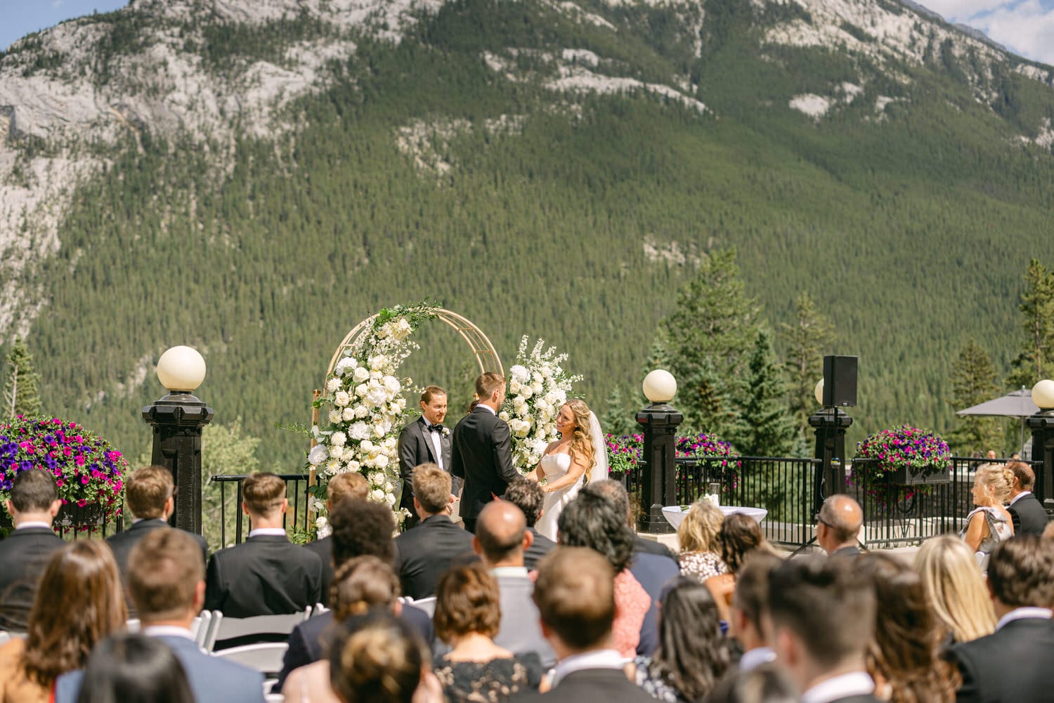 An outdoor wedding ceremony featuring a couple exchanging vows under a floral arch, surrounded by guests with mountains in the background.