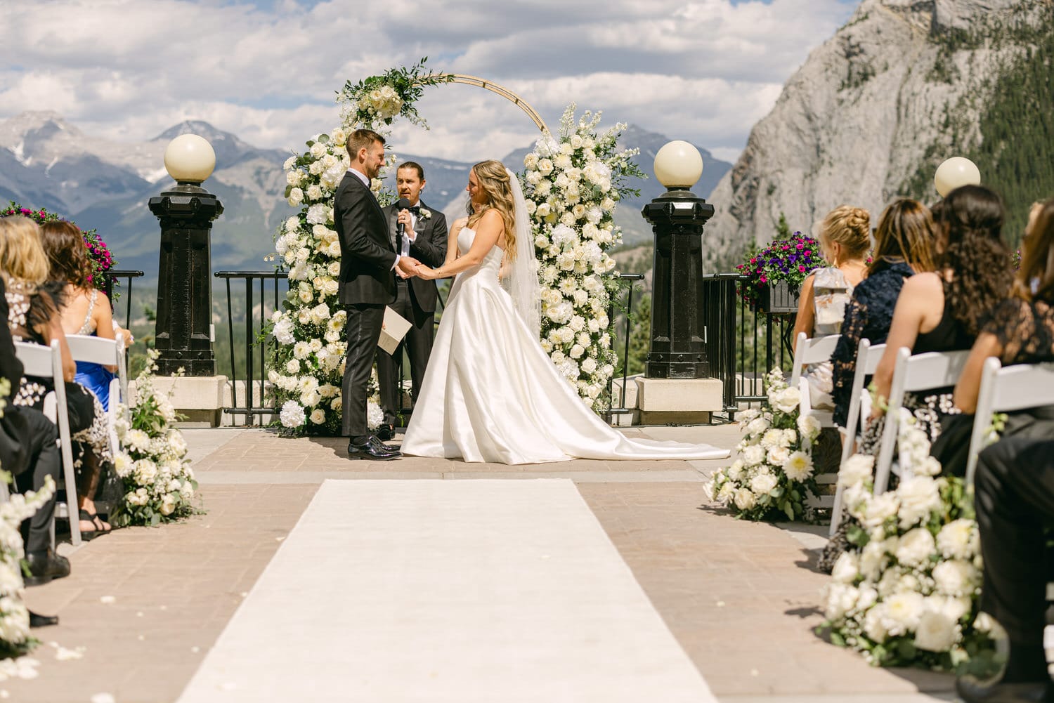 A bride and groom exchange vows during a romantic outdoor wedding ceremony surrounded by floral arrangements and scenic mountains in the background.