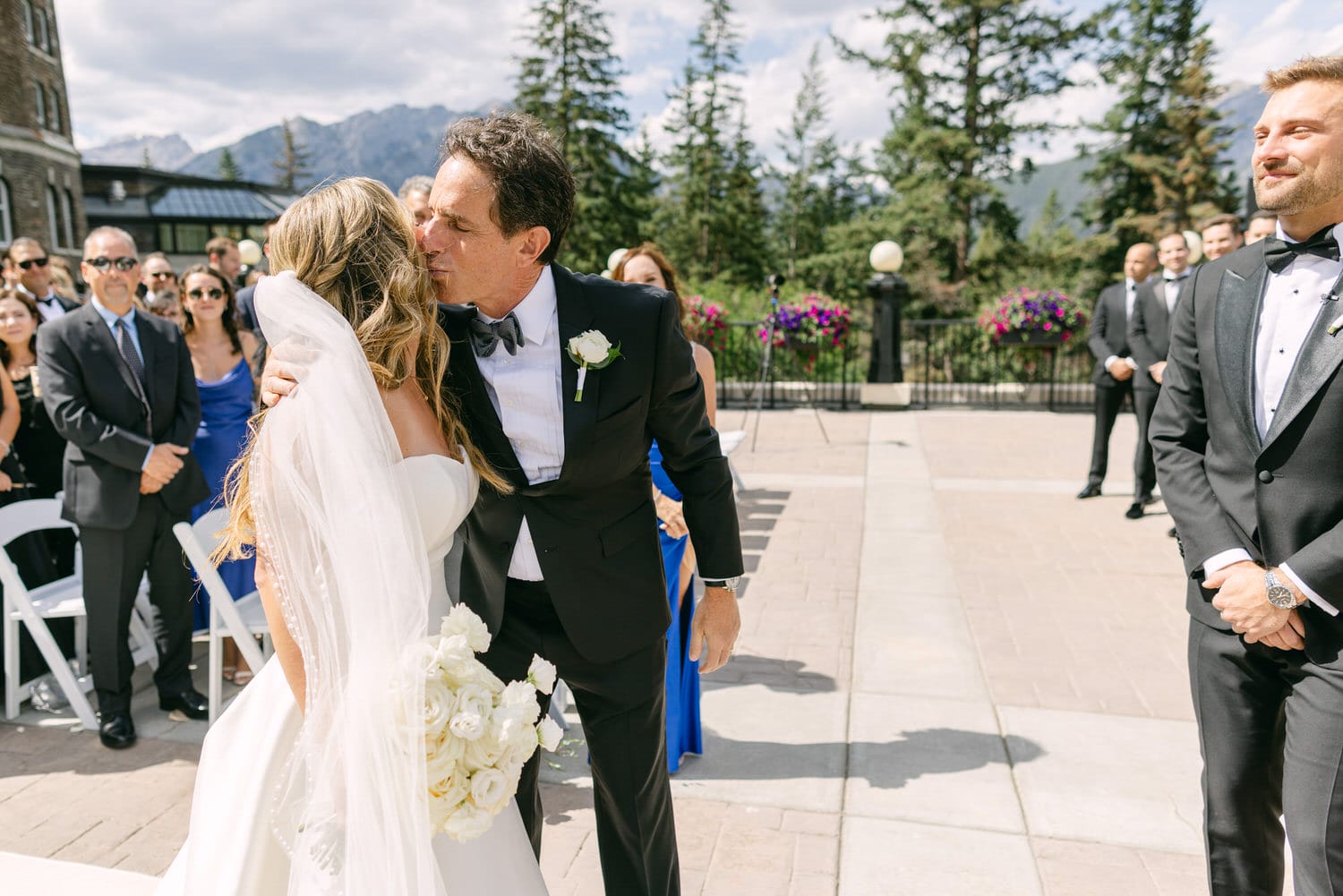 A joyous moment during a wedding ceremony as the bride receives a kiss from her father, surrounded by guests and a picturesque landscape.