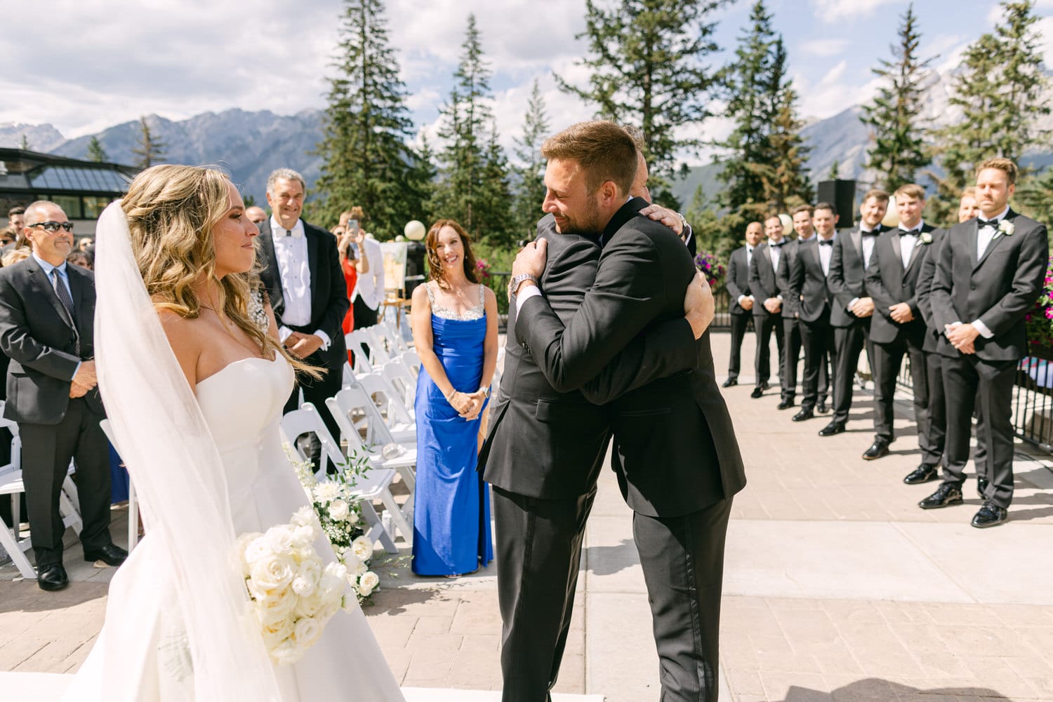 A groom shares a heartfelt embrace with a close friend during an outdoor wedding ceremony, surrounded by guests against a backdrop of mountains and trees.