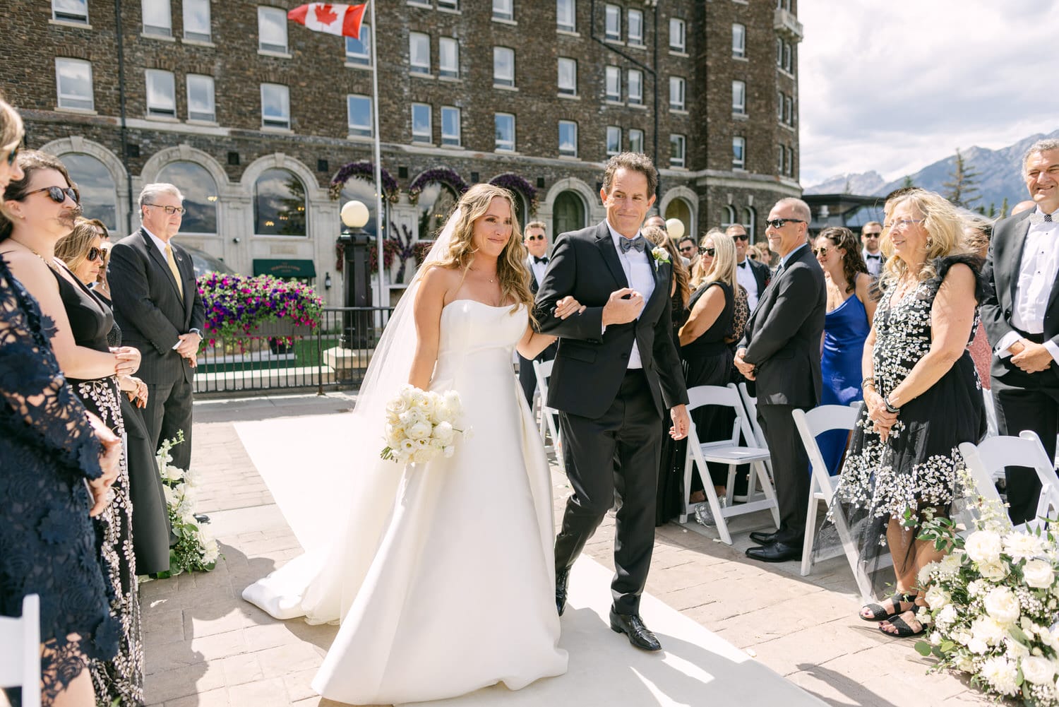 A bride in a white gown walks down the aisle with her father, surrounded by guests in formal attire at a picturesque outdoor wedding venue.