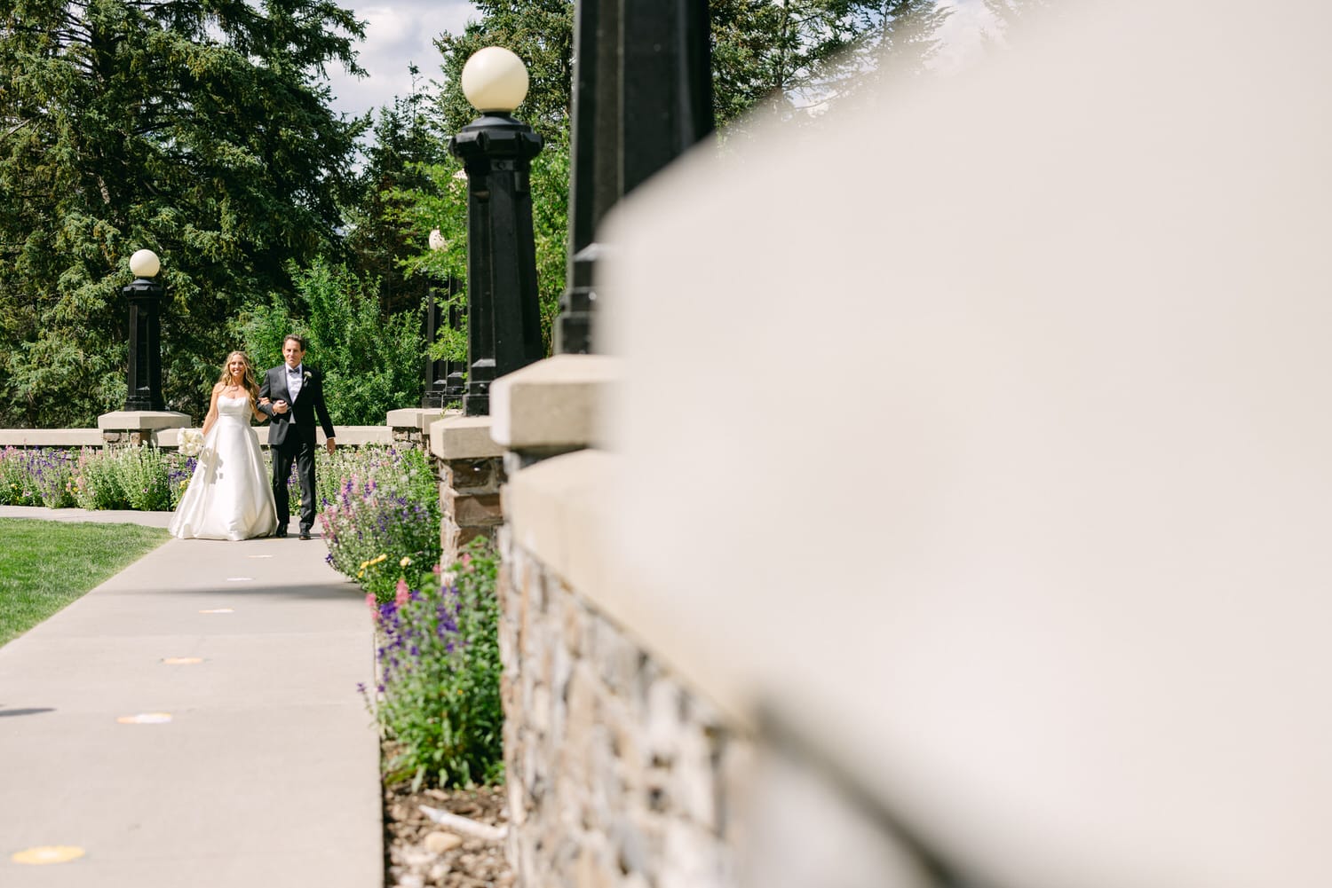 A bride and groom stroll down a scenic path surrounded by flowers and greenery, basking in the natural beauty on their special day.