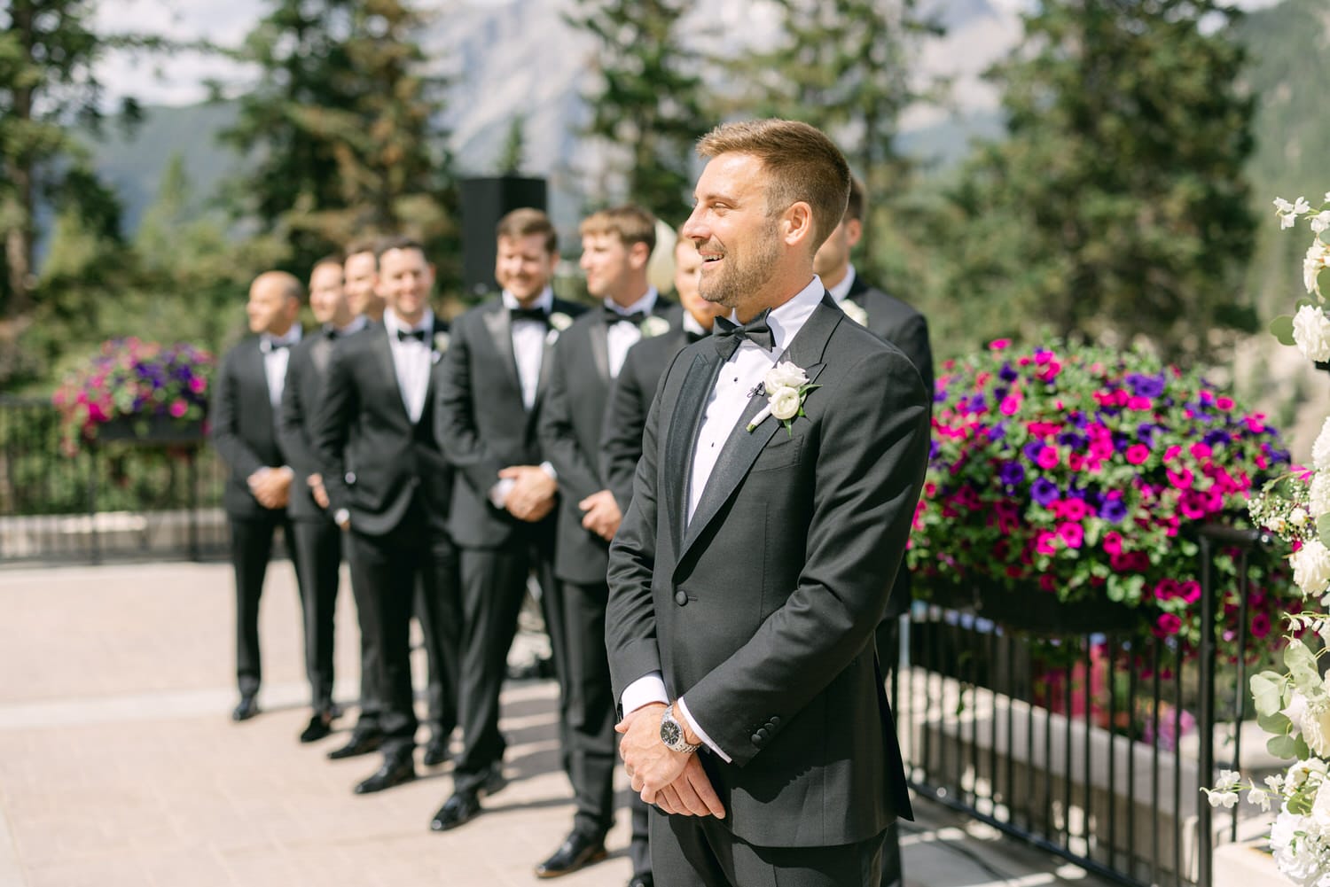 A groom in a black tuxedo stands with a smile, surrounded by groomsmen, against a backdrop of vibrant flowers and mountains during an outdoor wedding ceremony.