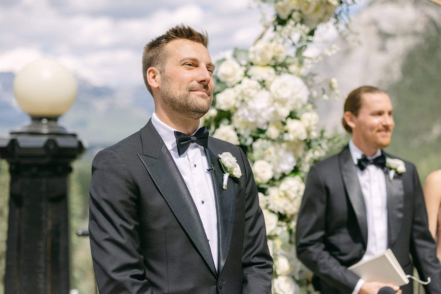 A well-dressed groom in a black tuxedo with a rose boutonniere stands smiling during an outdoor wedding ceremony, with floral decorations and guests in the background.