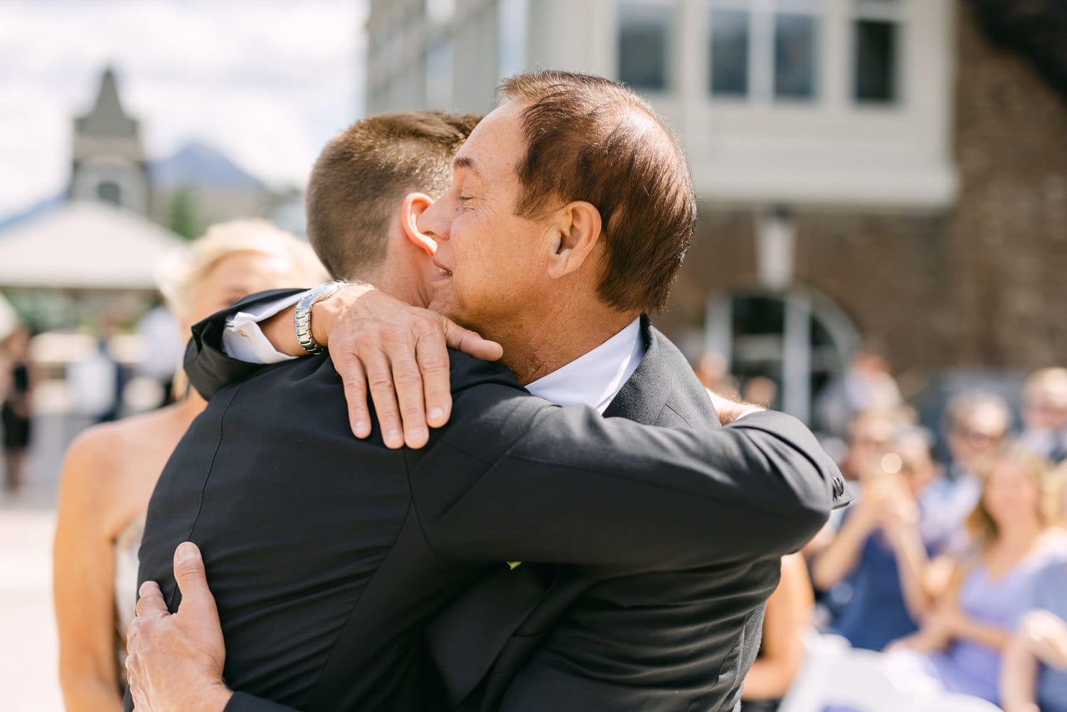 A heartfelt hug between two men during a wedding ceremony, surrounded by guests enjoying the moment.