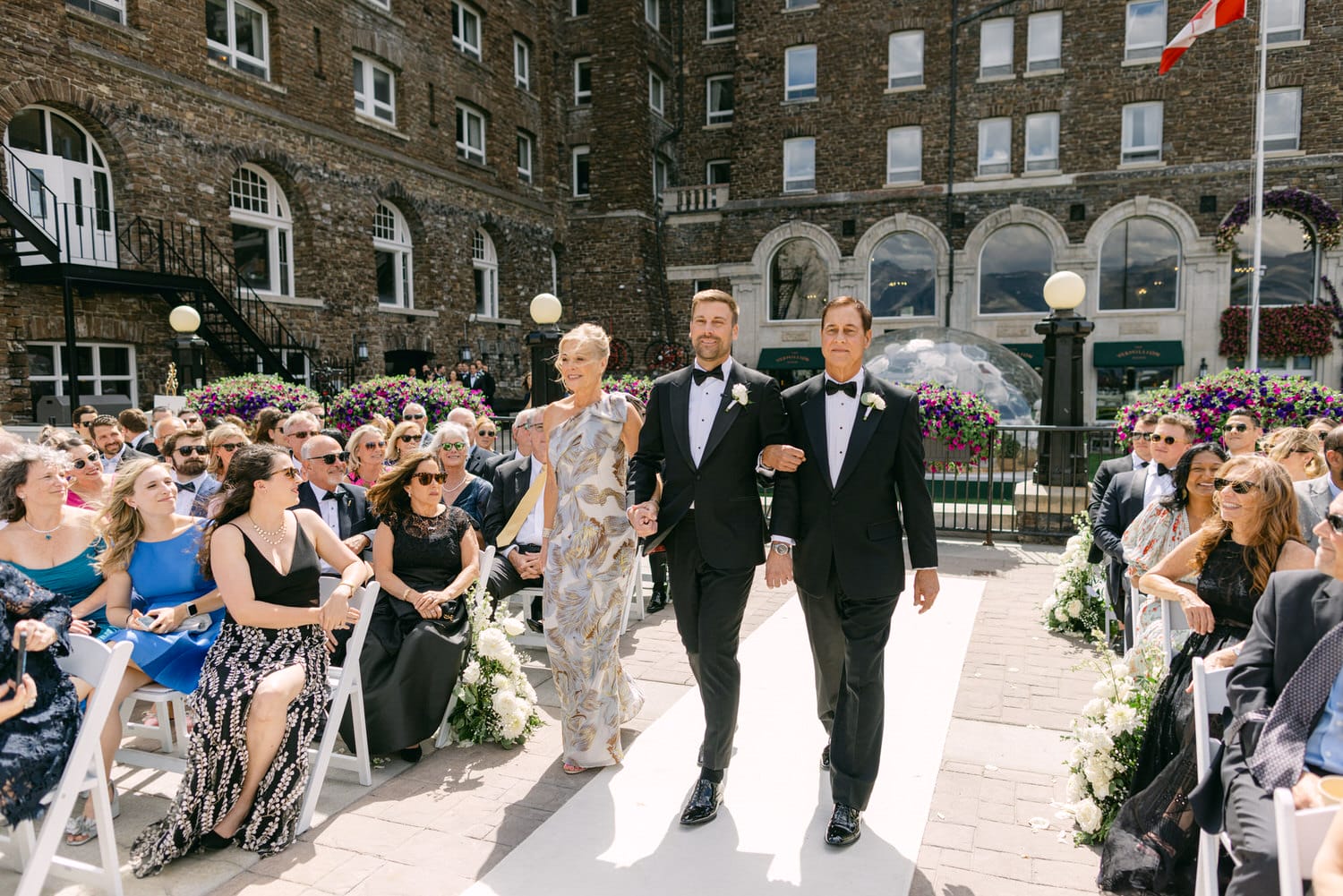 A groom walks down the aisle with his mother and father amid a floral backdrop, as guests look on, dressed in formal attire.