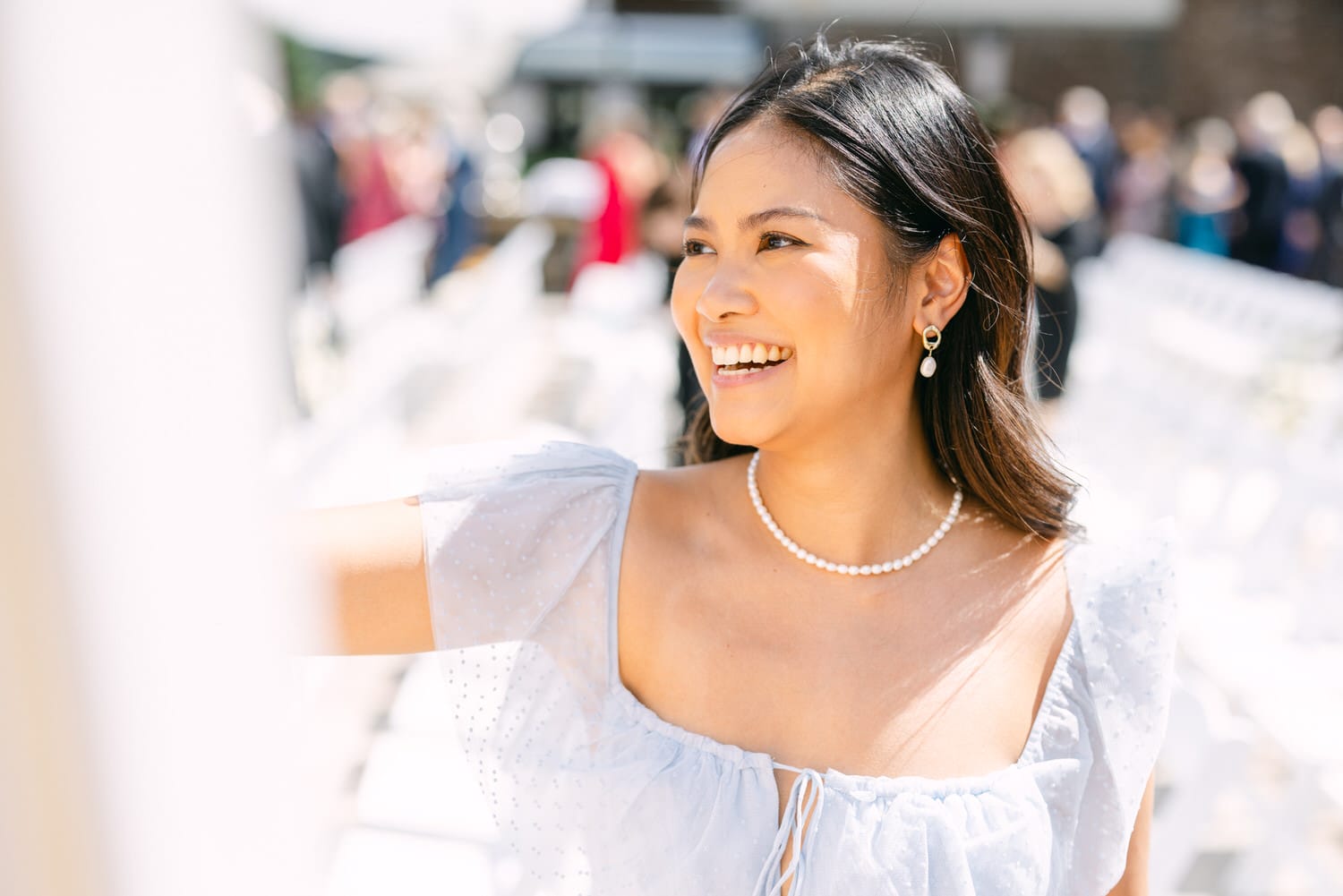 A smiling woman wearing a light blue dress and pearl accessories, capturing a joyful moment at an outdoor event.