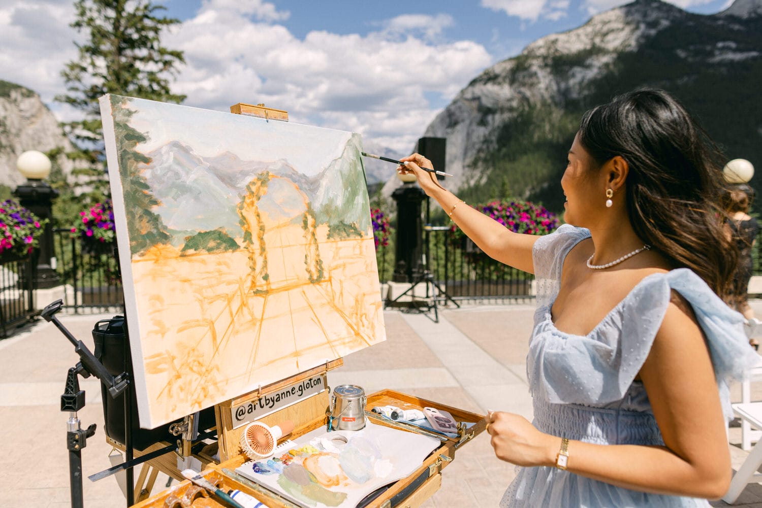 A woman painting a landscape on an easel, with mountains and a flower-filled backdrop.