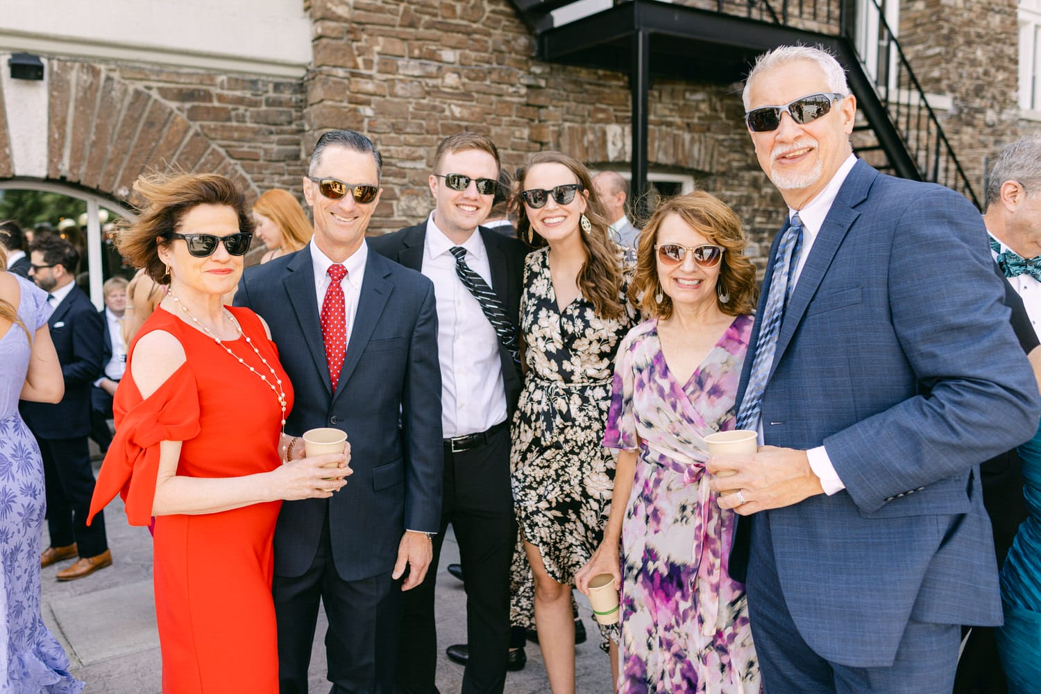 A group of six friends posing together at a sunny outdoor event, all wearing sunglasses and holding drinks, with a stone building in the background.