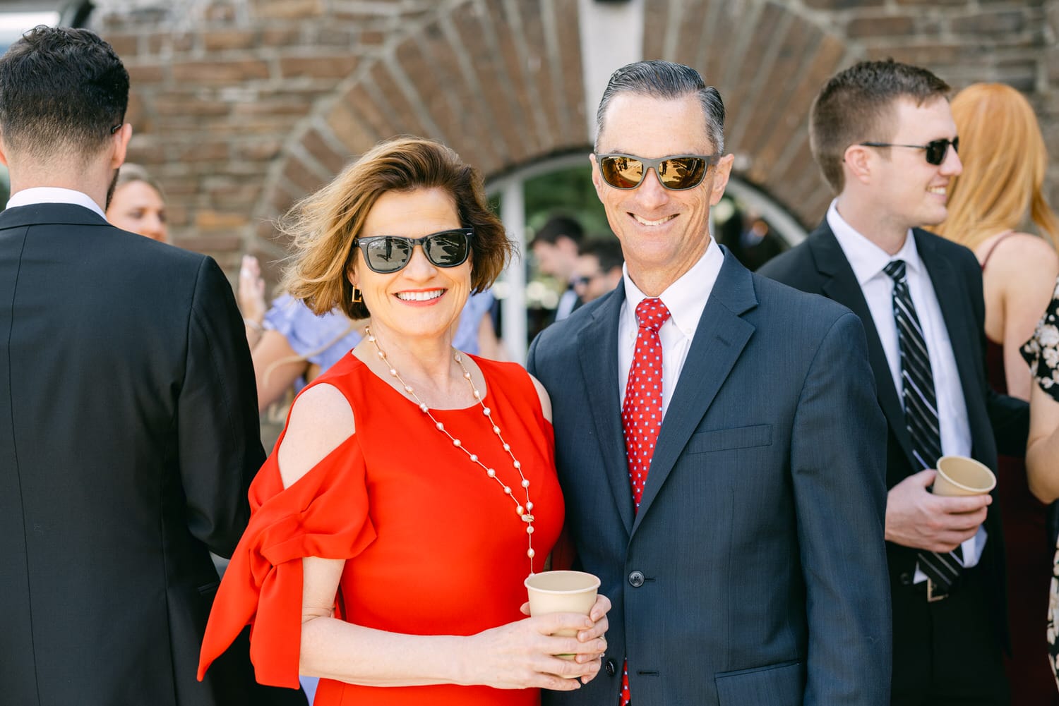 A smiling couple stands together at an outdoor event, the woman in a vibrant red dress and sunglasses, and the man in a suit with a polka-dot tie, both holding cups.